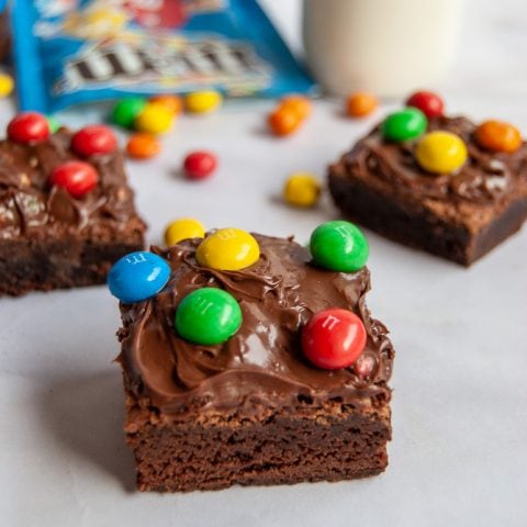 Chocolate Brownies topped with M&M spread and M&M candies on a white backdrop. A jar of M&M spread, a bottle of milk and a spilled packet of M&Ms are in the background