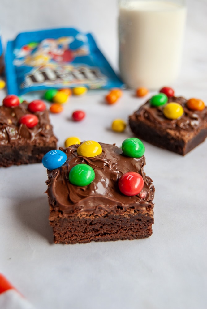 A brownie topped with M&M Spread and M&M Candies on a white background. A little bottle of milk, more brownies and a spilled packet of crispy M&Ms in the background.