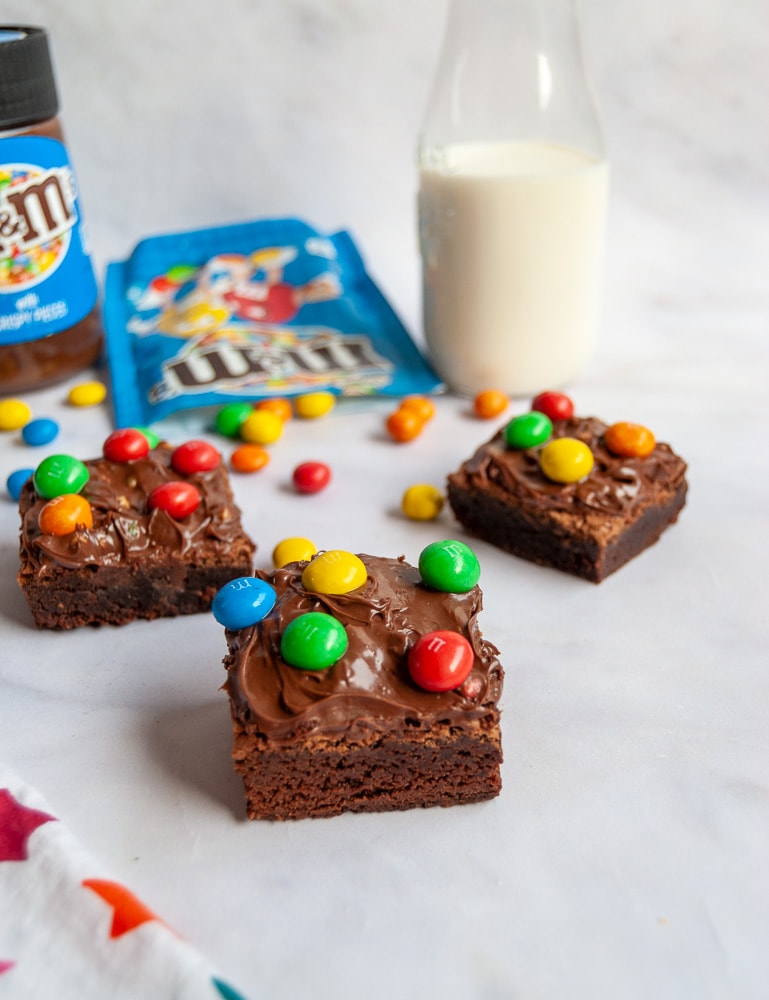 Chocolate Brownies topped with M&M spread and M&M candies on a white backdrop. A jar of M&M spread, a bottle of milk and a spilled packet of M&Ms are in the background