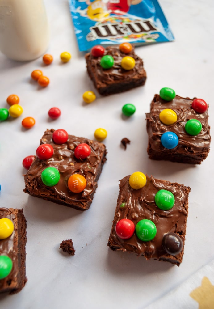 Chocolate Brownies topped with M&M spread and M&M candies on a white backdrop. A jar of M&M spread, a bottle of milk and a spilled packet of M&Ms are in the background