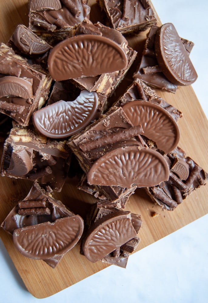 Pieces of no bake chocolate orange traybake with Matchmakers and Terry's chocolate orange segments on a wooden board.