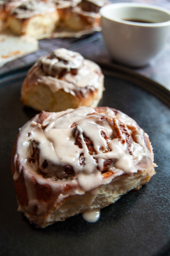 Two Cinnamon Rolls with a pumpkin puree filling and drizzled with a maple syrup glaze on a black plate. A white cup of coffee and more cinnamon rolls on a wire rack can be seen in the background.