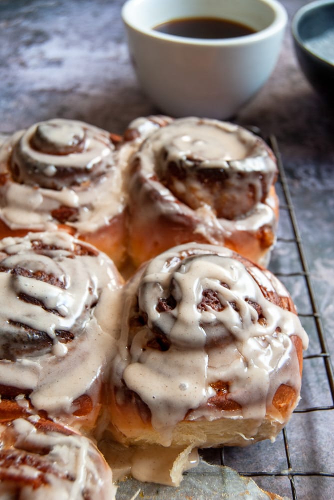 A close up picture of a batch of Pumpkin Cinnamon Rolls on a wire rack. A cup of coffee can be seen in the background.