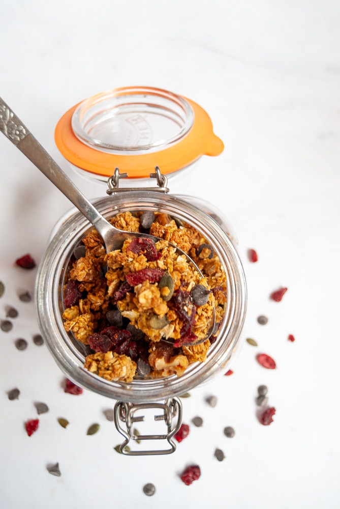 A closeup picture of an open jar of  granola with cranberries, pumpkin seeds and chocolate chips. A spoonful of granola is resting on top of the jar.