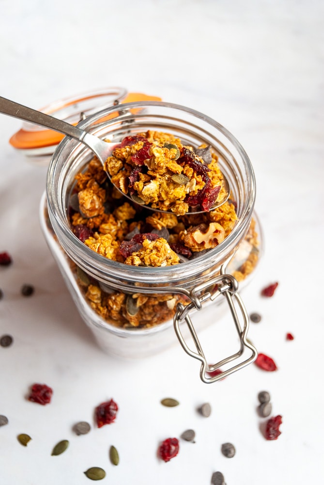 A jar of pumpkin spice chocolate chip granola on a white marbled background. A spoonful of granola is resting on the jar.