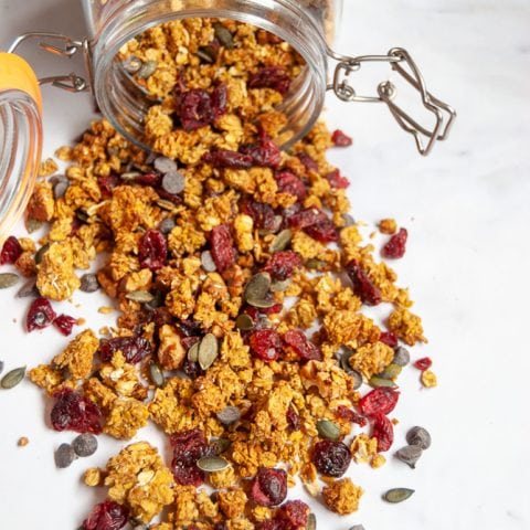 A spilled jar of Pumpkin spice chocolate chip granola with cranberries and pumpkin seeds on a marbled white background