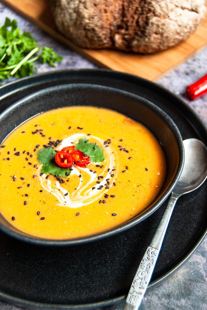 A black stoneware bowl of spicy sweet potato and lentil soup. A loaf of bread on a chopping board is in the background, A bunch of coriander and a red chilli are also in the picture.