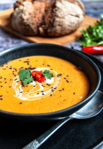 A black stoneware bowl of spiced sweet potato and lentil soup garnished with fresh coriander, chopped red chill and black sesame seeds.