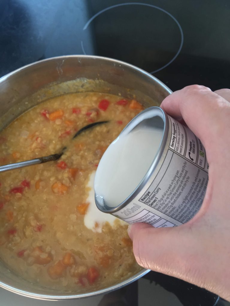 a small can of coconut milk being poured into a pan of spiced lentil soup.