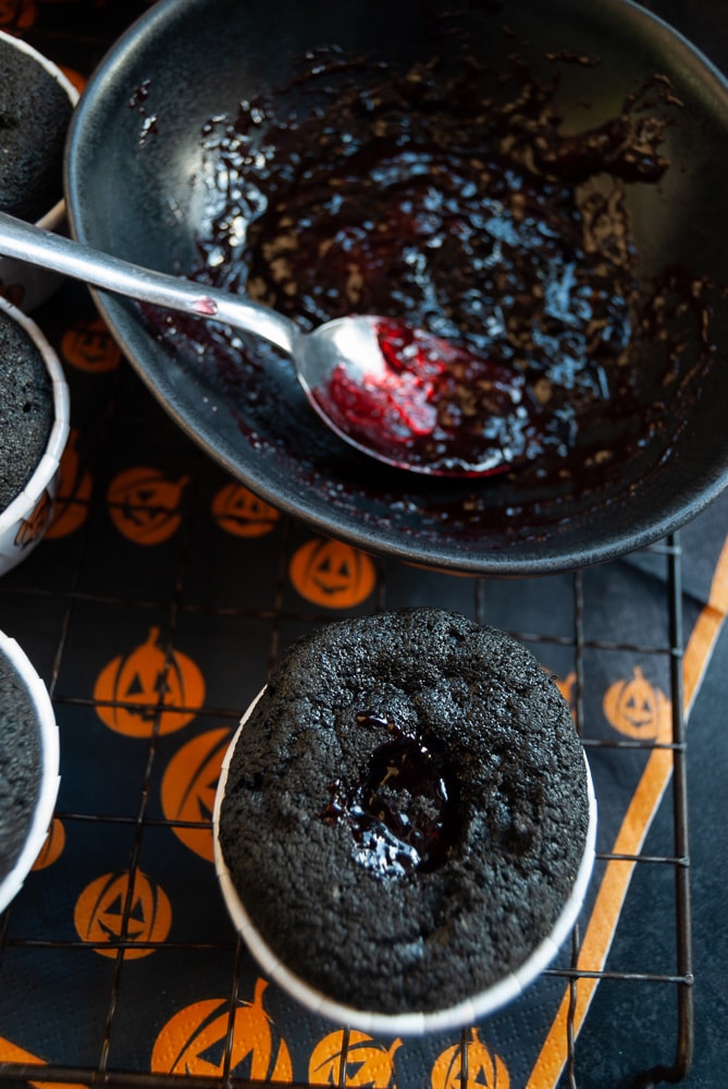 A black velvet cupcake filled with raspberry jam and a bowl of jam with a spoon.