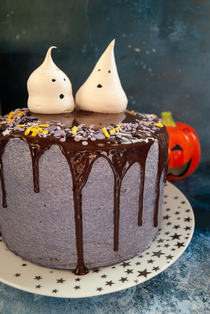 A photo of a Halloween chocolate cake decorated with purple buttercream, a chocolate ganache drip, Halloween sprinkles and Meringue ghosts. 