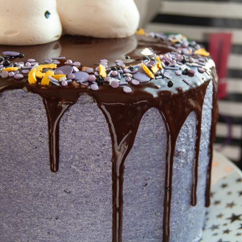 A close up photo of a Halloween drip cake decorated with purple buttercream, a chocolate ganache drip, purple and black Halloween sprinkles and Meringue Ghosts