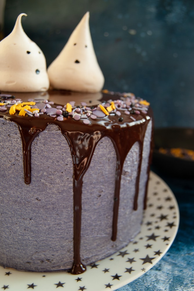 A Halloween cake decorated with purple buttercream and a chocolate ganache drip. Two meringue ghosts are sitting on top of the cake.