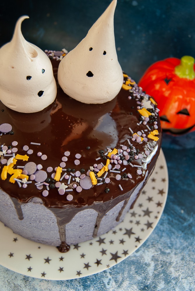 A chocolate Halloween cake decorated with purple buttercream, a chocolate drip running down the sides of the cake, purple and black sprinkles and meringue ghosts.