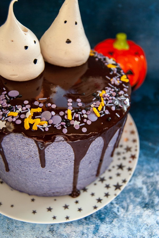 A chocolate Halloween cake decorated with purple buttercream, a chocolate ganache drip, Halloween spinkles and meringue ghosts.