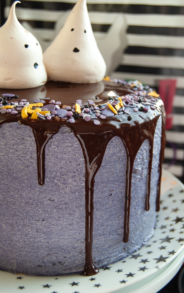 A close up photo of a Halloween cake decorated with purple buttercream, a chocolate ganache drip, purple and black Halloween sprinkles and Meringue Ghosts