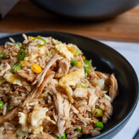 A bowl of leftover chicken fried rice with peas, sweetcorn and egg on a white background. a wok with more fried rice can partially be seen in the background