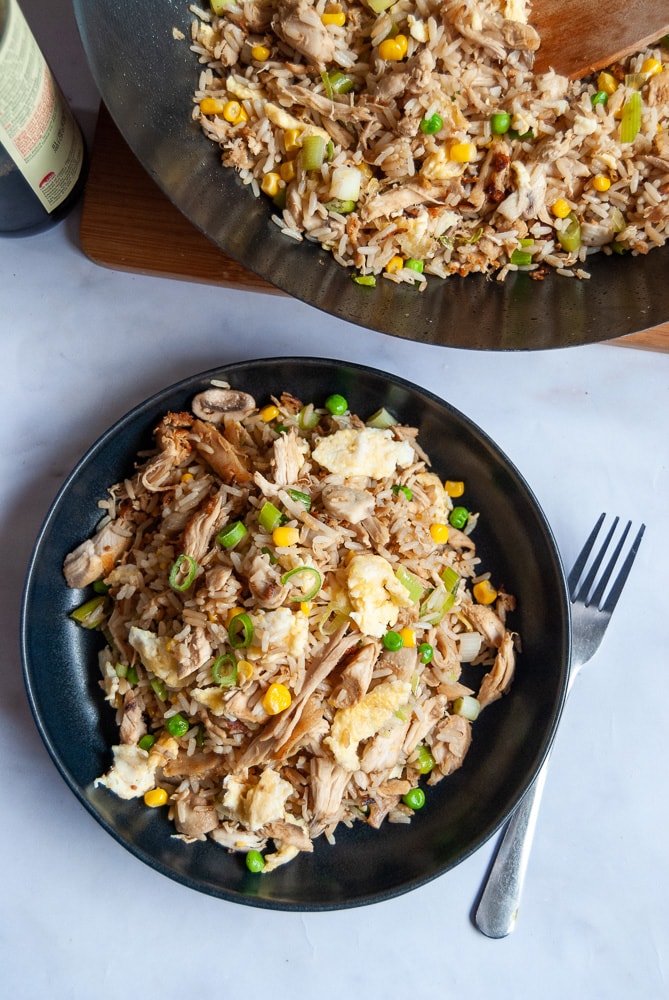 A black stoneware bowl of chicken fried rice. A wok with more fried rice can be seen in the top corner of the photo