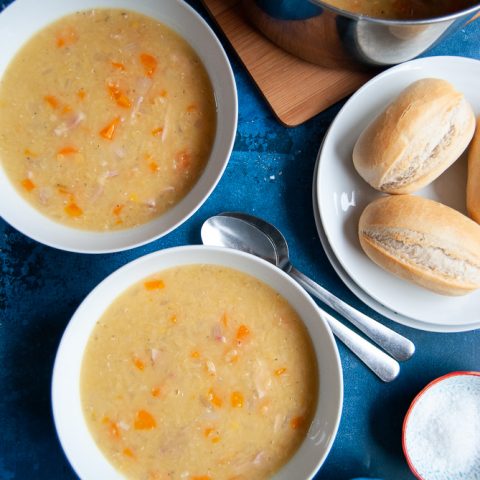 Two bowls of Lentil and Bacon Soup with a plate of bread rolls, salt and pepper and spoons on a dark blue background