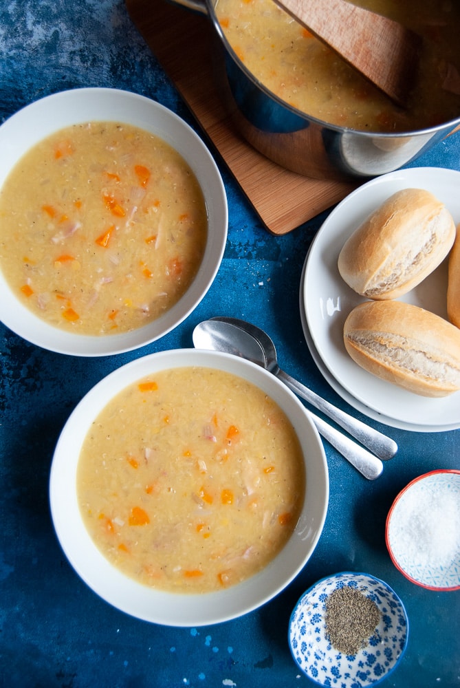 Two bowls of Lentil and Bacon Soup with a plate of bread rolls, salt and pepper and spoons on a dark blue background