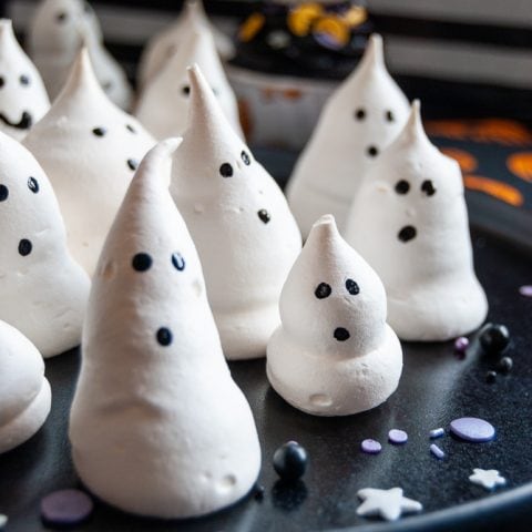LIttle ghosts made of piped meringue and black food colouring for eyes and a mouth on a black plate.