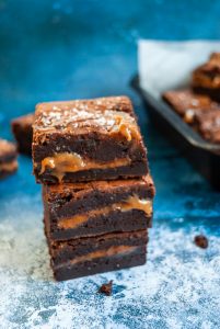 A stack of salted caramel brownies on a blue background. The brownies have an oozing salted caramel middle and are sprinkled with sea salt flakes