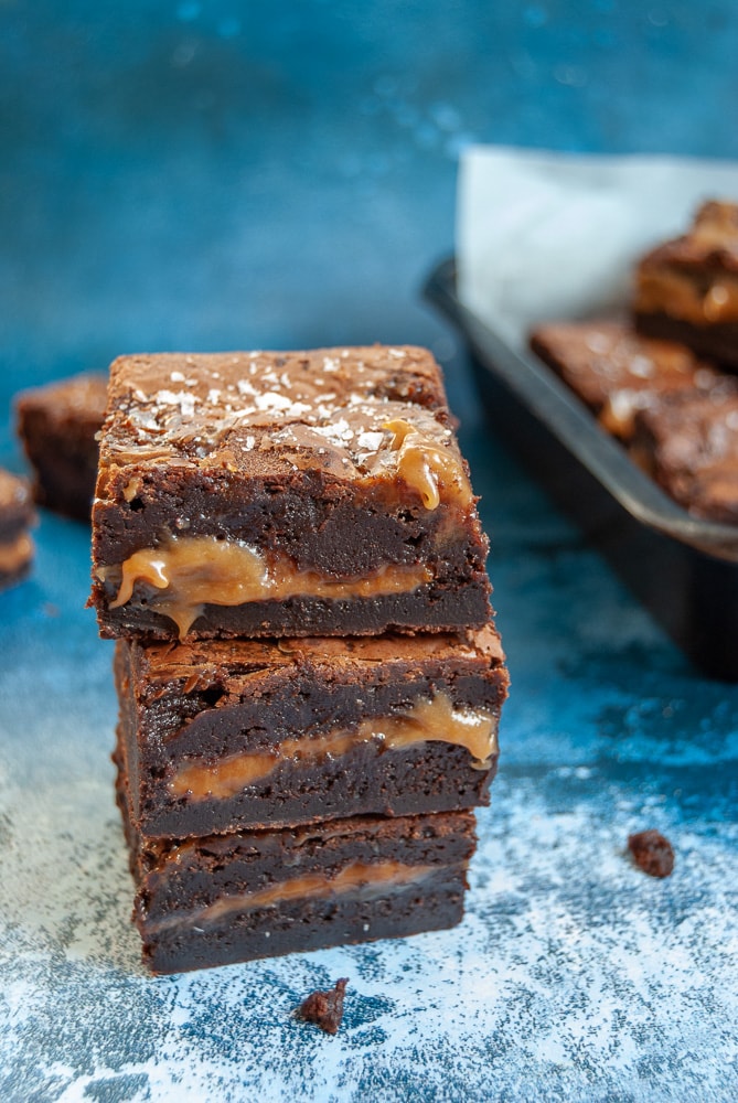 A stack of salted caramel brownies on a blue background. The brownies have an oozing salted caramel middle and are sprinkled with sea salt flakes