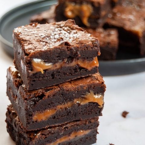 A stack of chocolate brownies with a salted caramel middle. A plate of brownies can be seen in the background