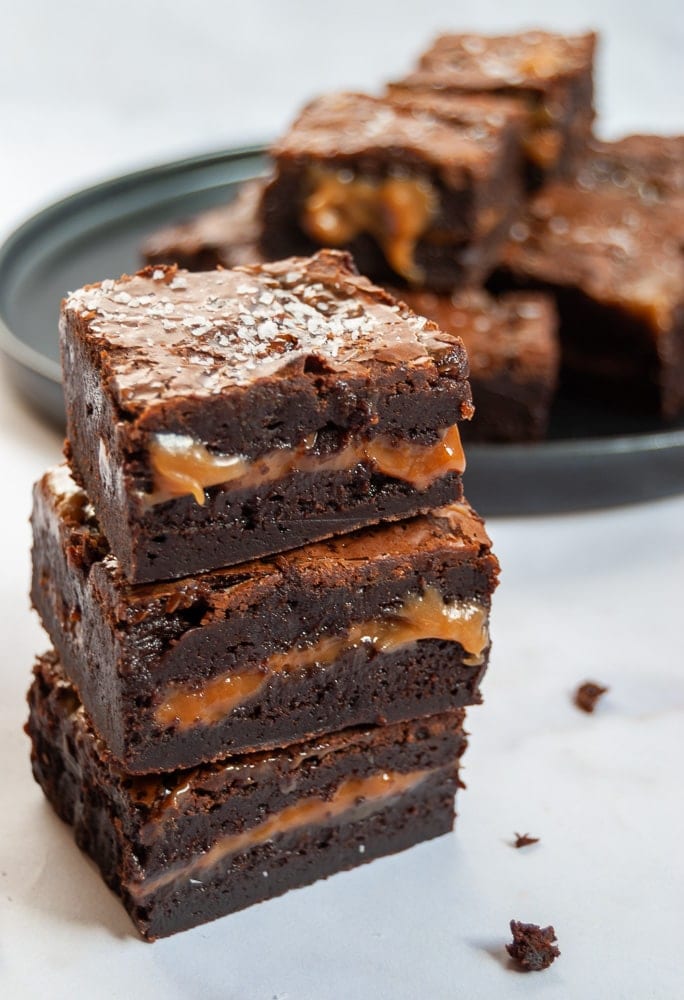 A stack of chocolate brownies with a salted caramel centre. A black plate of brownies can be seen in the background