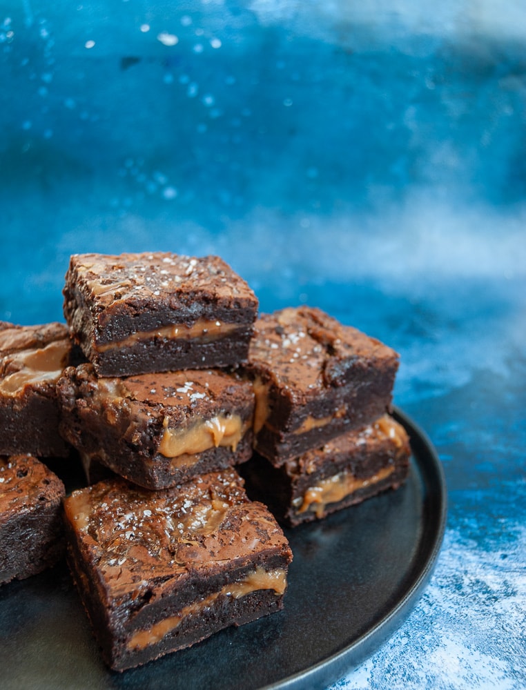 Chocolate brownies with a salted caramel middle piled up on a black plate. 