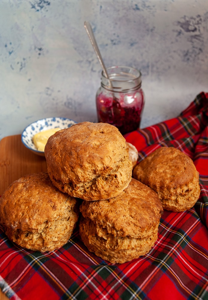 A pile of scones on a red tartan cloth.  A little bowl of butter and a jar of jam can be seen in the background