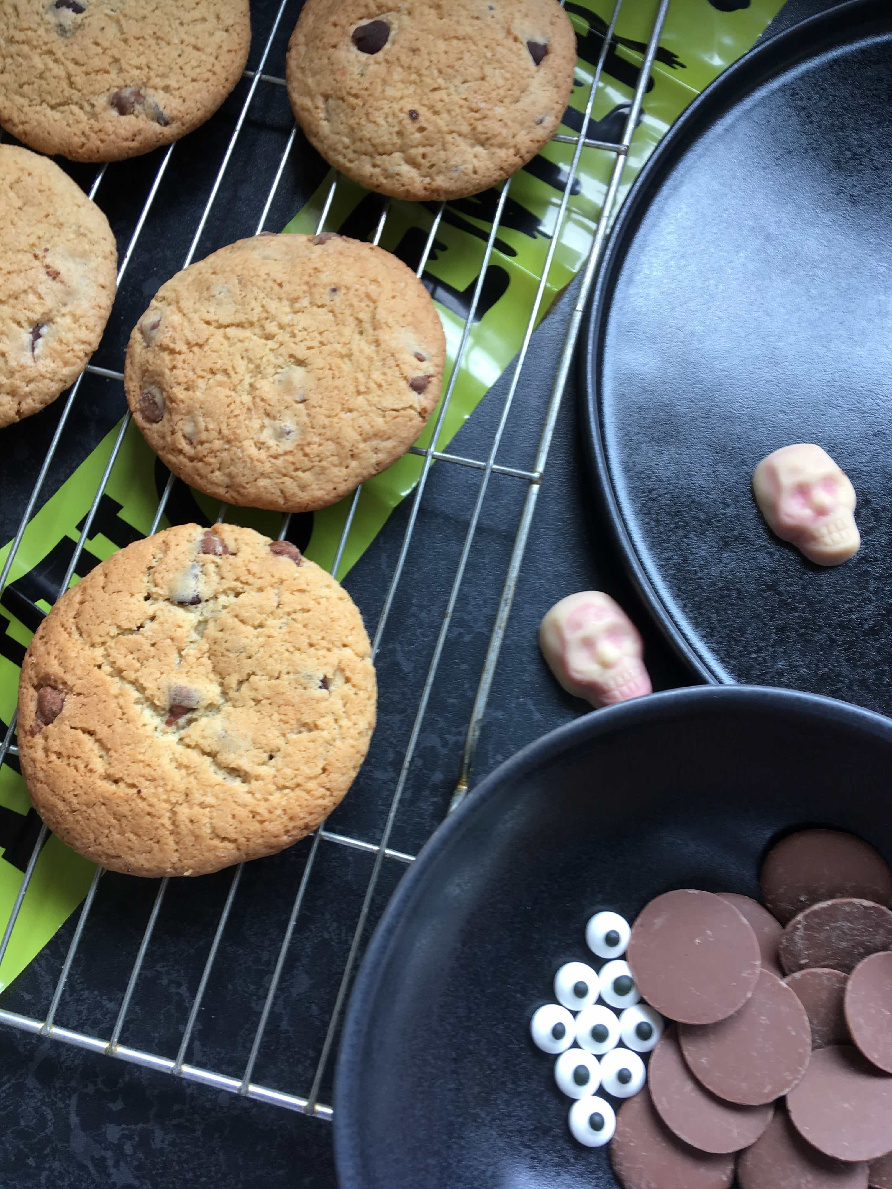 A Wire rack of chocolate chip cookies with a bowl of candy edible eyes and chocolate buttons