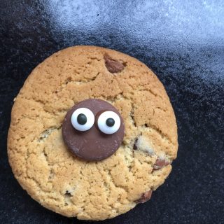 A chocolate chip cookie decorated with a chocolate button and candy eyes 