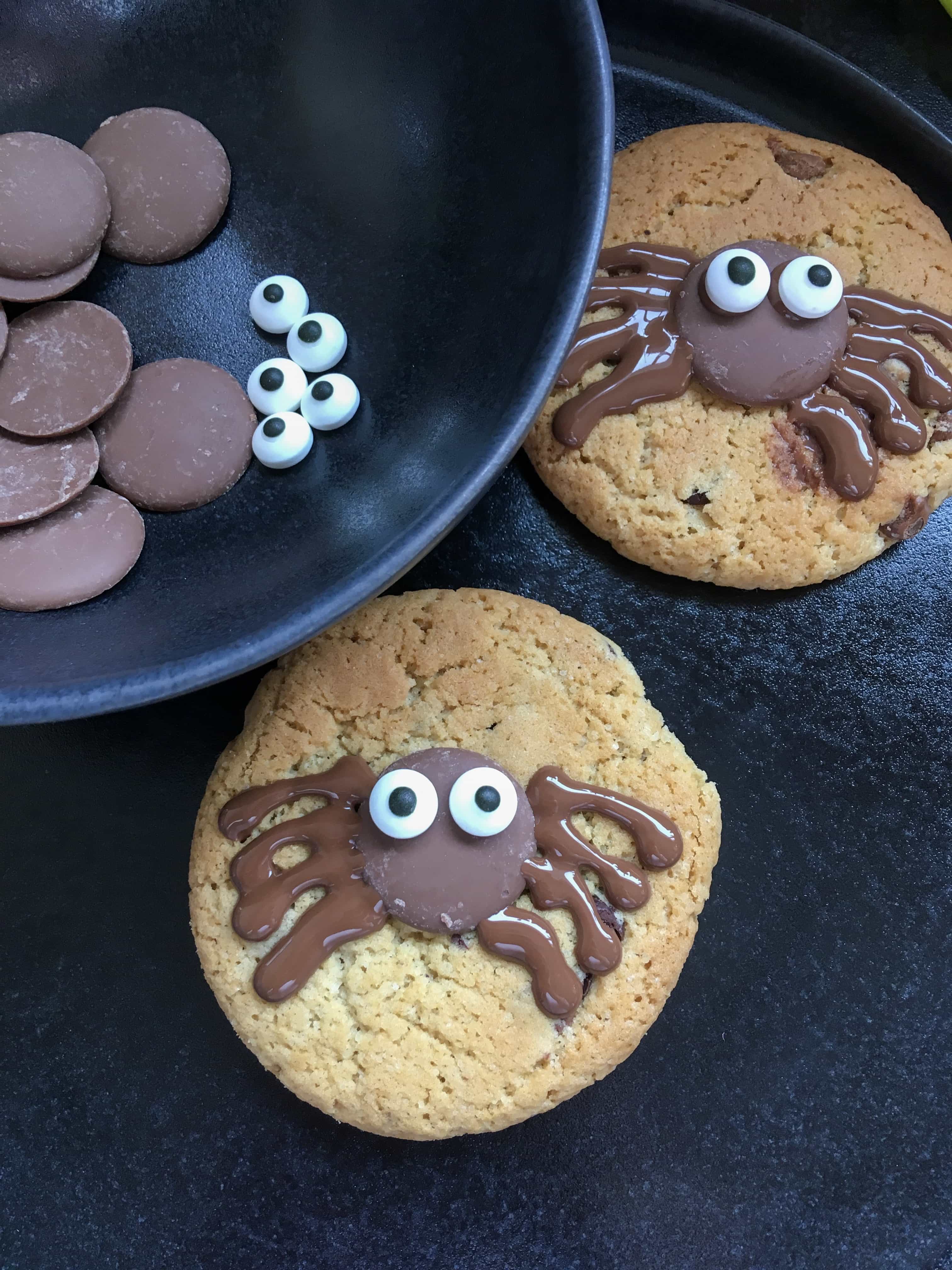 Two Spooky spider cookies sitting on a black plate - chocolate chip cookies decorated with a chocolate button, candy eyes and melted chocolate legs