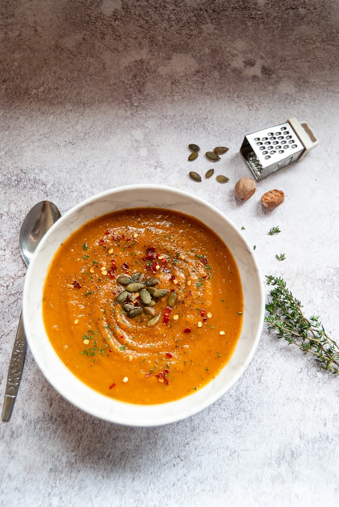 A white marbled bowl of butternut squash soup garnished with fresh thyme, pumpkin seeds and red chilli flakes