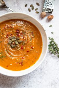 A flat lay photo of a bowl of butternut squash and sweet potato soup garnished with pumpkin seeds, chilli flakes and thyme.