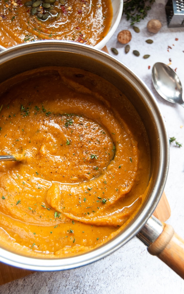 A pan and white bowl of butternut squash and sweet potato soup, garnished with herbs, spices and pumpkin seeds on a wooden chopping board.