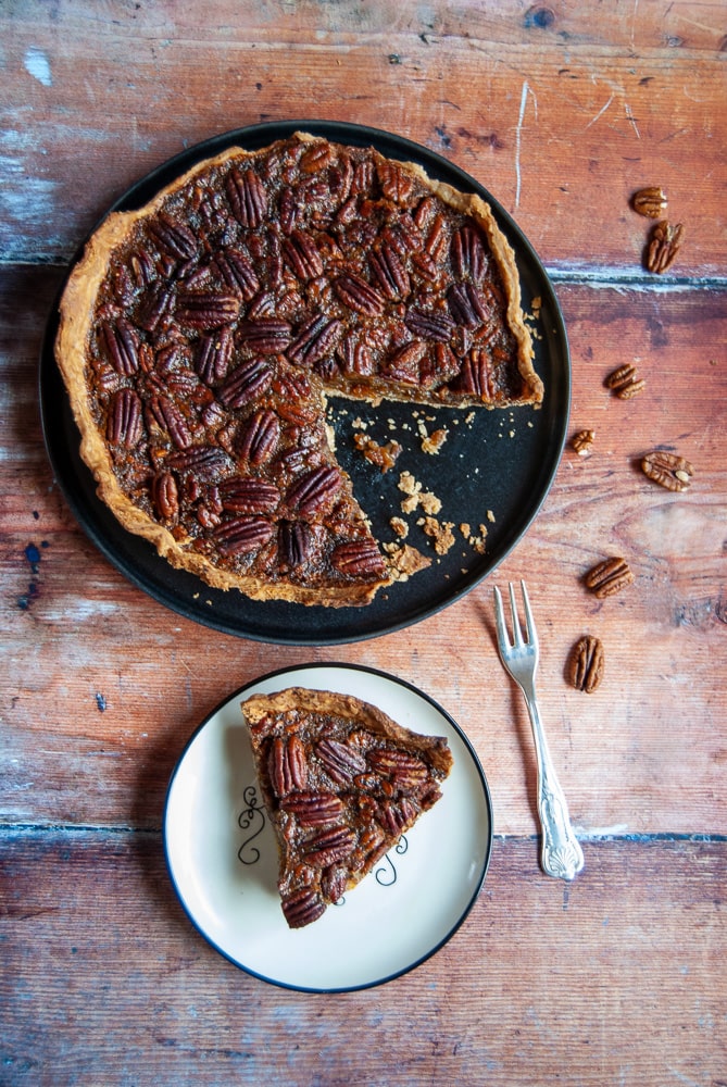 a slice of pecan pie on a white plate and a large pecan pie on a black plate with a silver fork.