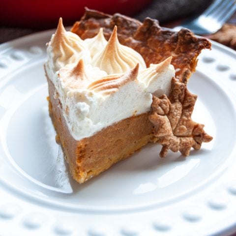 A slice of pumpkin pie topped with meringue on a white plate with a maple leaf biscuit