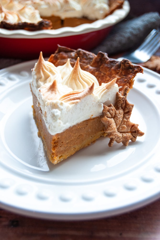 A slice of pumpkin pie topped with meringue on a white plate with a maple leaf biscuit