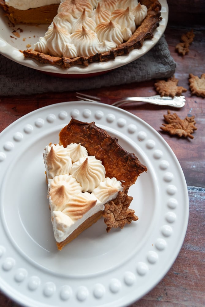 A slice of pumpkin pie topped with meringue on a white Plate/wooden backdrop. 