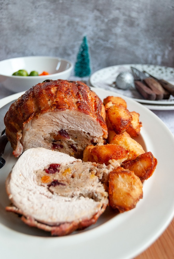 A rolled turkey bread stuffed with sausagemeat,, cranberry and apricot stuffing with roast potatoes in a white plate. 