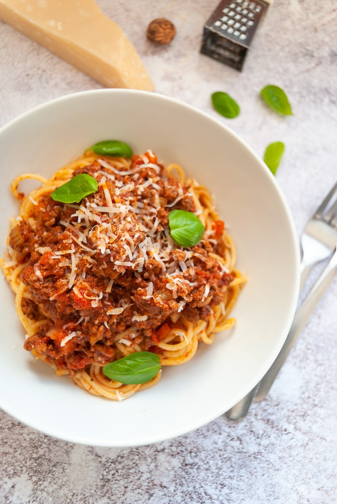 A close up photo of a bowl of slow cooked bolognese sauce with spaghetti on a white and grey background.