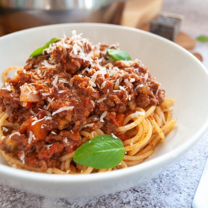 A bowl of spaghetti bolognese with grated parmesan and fresh basil leaves on a white and grey background