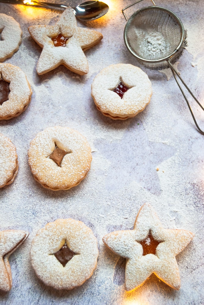 Christmas Linzer Cookies on a grey and white background dusted with icing sugar. Fairy lights and a small metal sieve are also in the picture.