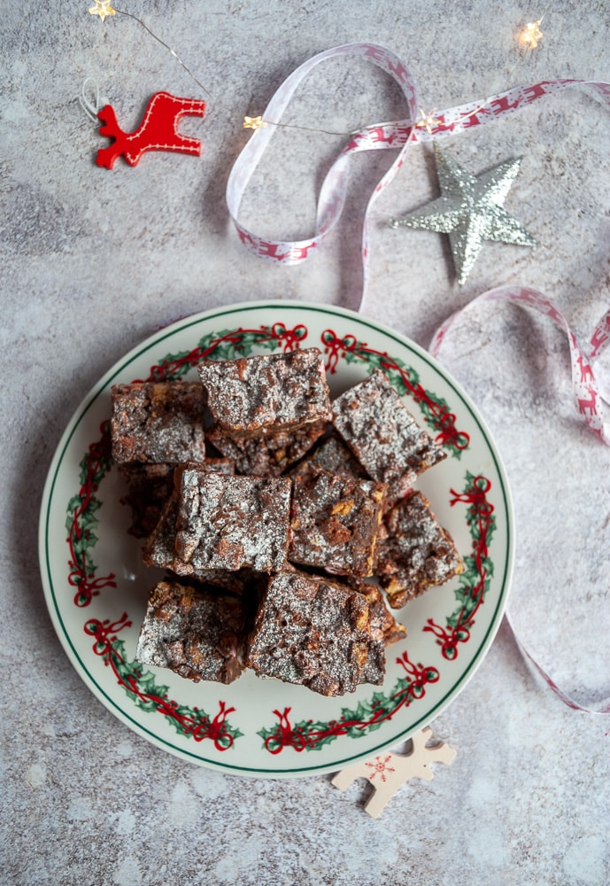 A plate of rocky road dusted with icing sugar, red and white reindeer and red and white christmas ribbon on a grey backdrop