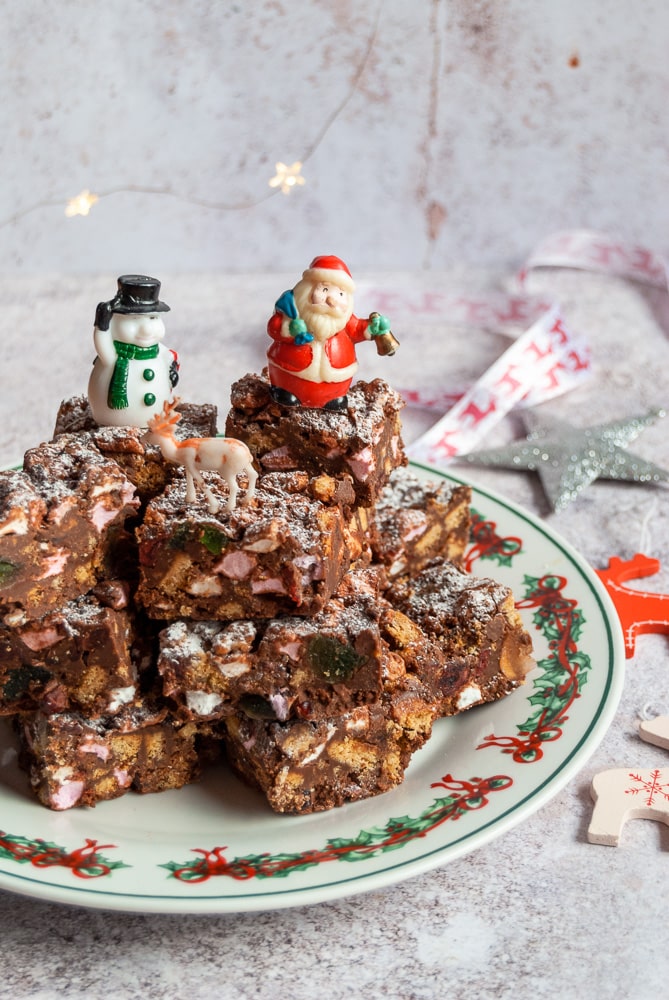 rocky road bars piled up on a Christmas plate with santa, snowman and reindeer figures. Fairy lights can be seen in the background.