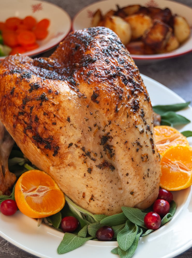 A Roast Turkey Crown on a white plate with sage leaves, cranberries and halved oranges.