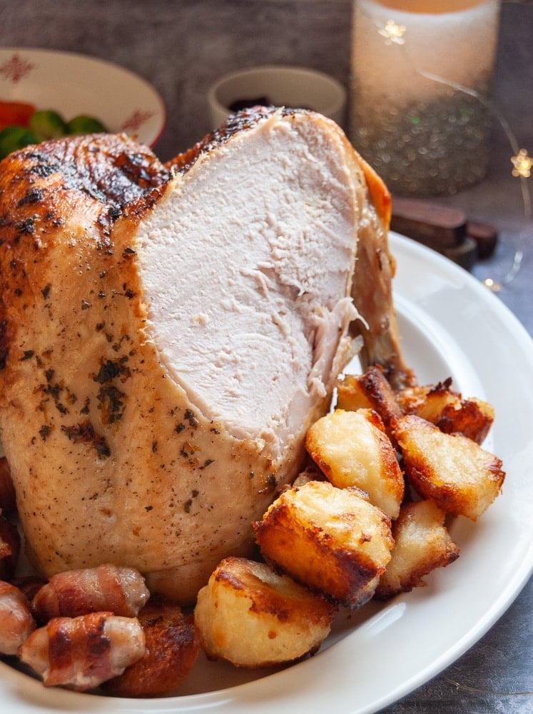 A roast turkey crown on a white plate with roast potatoes and bacon wrapped sausages.
