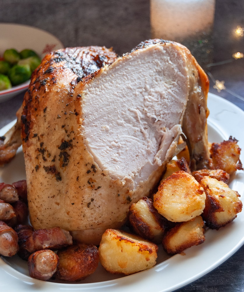 A roast turkey crown with a slice cut out on a white plate with roast potates and bacon wrapped sausages